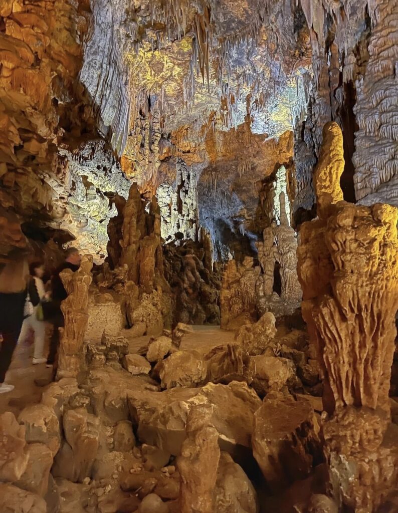 Interno di una grotta con stalattiti e stalagmiti illuminate da luci calde. Persone che esplorano il luogo.